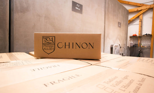 Wine bottle box with Chinon written on the side sitting on top of all the other boxes of wine in front of large cement tanks