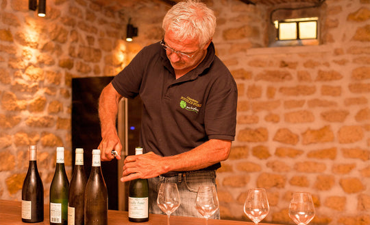 Regis Minet opening a bottle of his white wine in a large stone building. glasses and bottles of wine populate the table